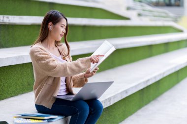 Asyalı kız öğrenci üniversite alanında sınavlara hazırlanıyor. İnternete bağlı cep telefonu ya da dizüstü bilgisayarla iletişim kurun. bir çok bilgi kaynağıyla