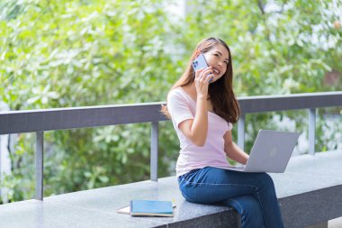 Asyalı kız öğrenci üniversite alanında sınavlara hazırlanıyor. İnternete bağlı cep telefonu ya da dizüstü bilgisayarla iletişim kurun. bir çok bilgi kaynağıyla