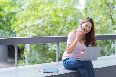 Asyalı kız öğrenci üniversite alanında sınavlara hazırlanıyor. İnternete bağlı cep telefonu ya da dizüstü bilgisayarla iletişim kurun. bir çok bilgi kaynağıyla