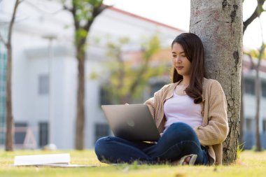 Asyalı kız öğrenci üniversite alanında sınavlara hazırlanıyor. İnternete bağlı cep telefonu ya da dizüstü bilgisayarla iletişim kurun. bir çok bilgi kaynağıyla