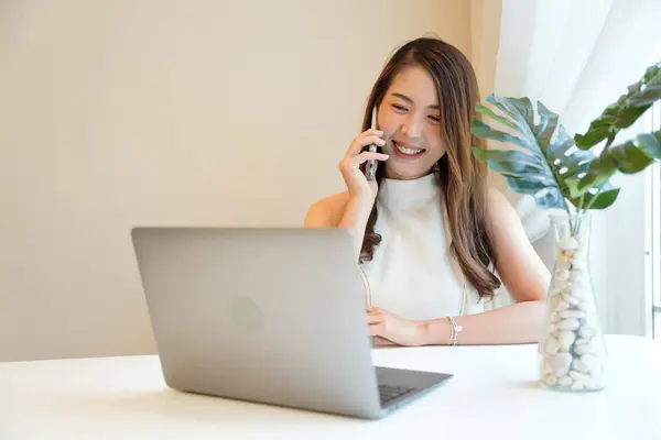 Asian woman with curly hair is working chatting with clients or colleagues via communication channels, telephone, internet with her laptop and mobile phone.