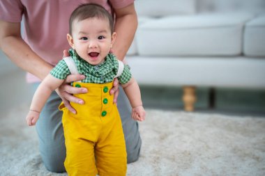 Bir adam bir bebek tutuyor ve gülümsüyor. Bebek sarı bir kıyafet giyiyor. Sahne mutlu ve neşeli.