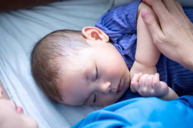 Bir kadın göğsünde bir bebekle kanepede yatıyor. Bebek uyuyor ve kadın onu kucağında tutuyor. Kanepe kahverengidir ve oda loş aydınlıktır.