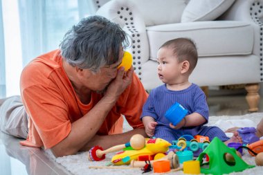 Bir adam ve bir bebek oyuncaklarla çevrili şekilde yerde yatıyorlar. Adam sarı bir oyuncak tutuyor ve bebek mavi bir oyuncak tutuyor. Sahne neşeli ve şen şakrak.