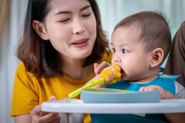 Bir kadın yüksek sandalyede bir bebeği besler. Bebek önlük takıyor ve yemek yiyor. Sahne sıcak ve sevgi dolu, kadın ve bebek birlikte özel bir an paylaşıyorlar.