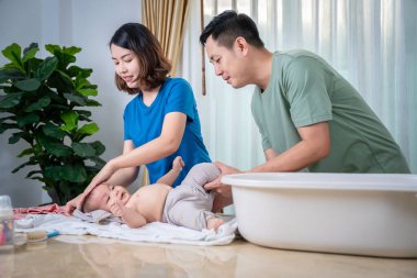 Anne ve baba, bebeklerini küvette yıkamak için birbirlerine yardım ediyorlar. Resimde herkes mutlu görünüyor..