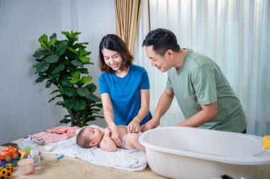 Anne ve baba, bebeklerini küvette yıkamak için birbirlerine yardım ediyorlar. Resimde herkes mutlu görünüyor..