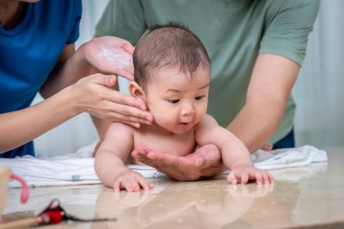 Bir bebek iki yetişkin tarafından nazikçe temizleniyor. Biri ellerine pudra sürerken, diğeri havlunun üzerinde yatan bebeği destekliyor. Bebek önlerindeki bir şeye odaklanmış görünüyor, tuhaf bir ifadeyle..