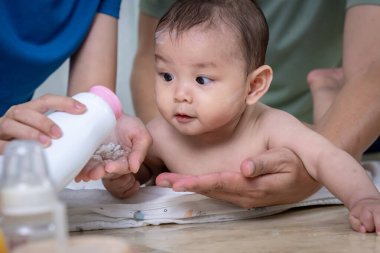 Bir bebek iki yetişkin tarafından nazikçe temizleniyor. Biri ellerine pudra sürerken, diğeri havlunun üzerinde yatan bebeği destekliyor. Bebek önlerindeki bir şeye odaklanmış görünüyor, tuhaf bir ifadeyle..