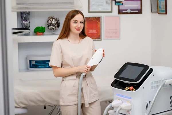 Stock image Beautician holding ultrasound device for face and skin lifting procedure and hair removal. Young woman cosmetologist using modern cosmetology equipment in clinic.