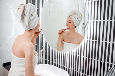 Banyo aynasının önünde dişlerini fırçalayan beyaz havlu giymiş güzel bir genç kadının portresi. Sabah rutin güzellik prosedürü