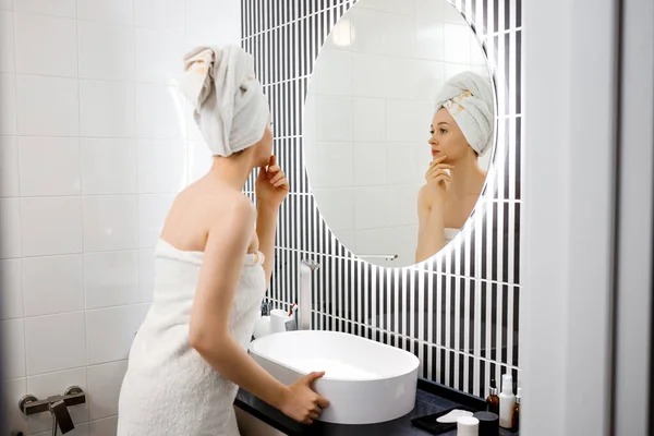 穿着毛巾的迷人的年轻女人在洗澡后看着镜子站在浴室里 美容美发概念 — 图库照片