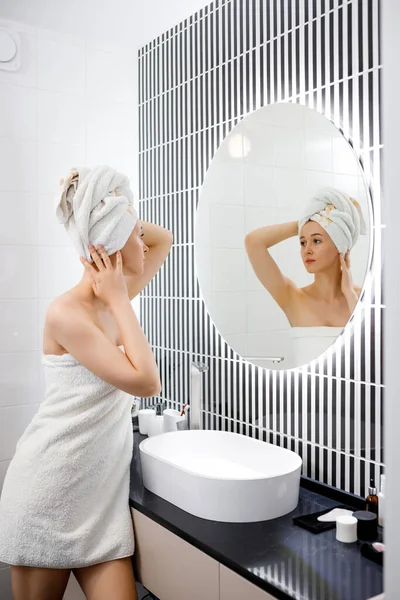 穿着毛巾的迷人的年轻女人在洗澡后看着镜子站在浴室里 美容美发概念 — 图库照片