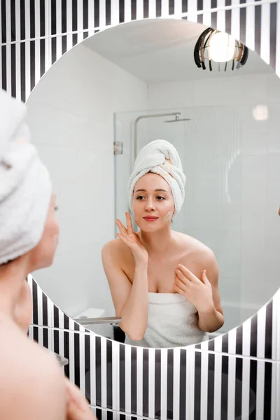 Young woman applying anti-wrinkle cream standing behind mirror in home bathroom. Cosmetology and beauty procedure. Skin care after cleansing