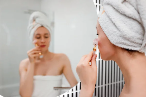 シャワーの後に白いタオルを身に着けて完璧な肌を持つ美しい女性 バスルームで天然クォーツ石と玉顔ローラーを使用して顔のマッサージを作ります 天然化粧品のコンセプト しわの平滑化 — ストック写真