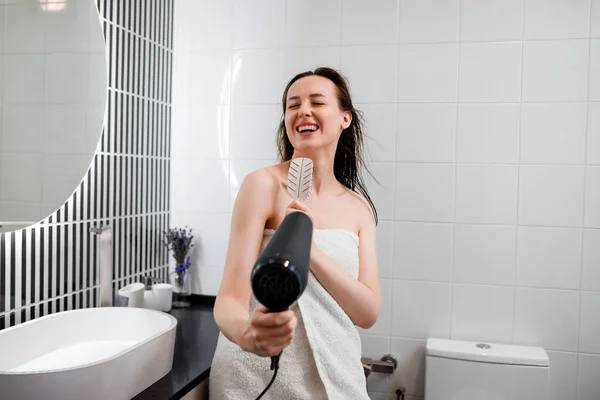 朝のシャワーの後 バスルームでヘアドライヤーでボリュームのある髪を乾かしながら 楽しさ ダンス 歌を持つ幸せな笑顔の女性 — ストック写真