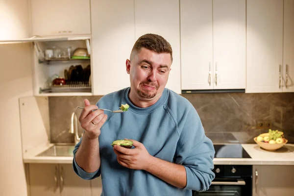 Fat and funny man eating avocado while standing in modern home kitchen. Slimming and healthy lifestyle concept