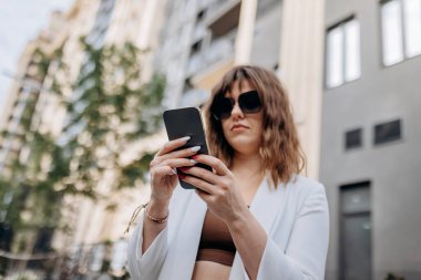 Smiling businesswoman in white suit using phone during walking in city with modern architecture