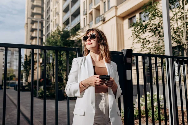 Smiling businesswoman in white suit using phone during walking in city with modern architecture and looking at side