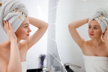 Havlu giymiş çekici genç bir kadın duş aldıktan sonra banyoda duran aynaya bakıyor. Güzellik ve Cilt Konsepti