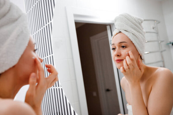 Young woman applying anti-wrinkle cream standing behind mirror in home bathroom. Cosmetology and beauty procedure. Skin care after cleansing