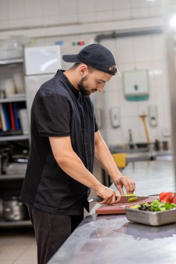 Professional smiling chef in uniform cut fresh vegetables in kitchen. Cook man cooking delicious garnish for clients in restaurant. Culinary concept