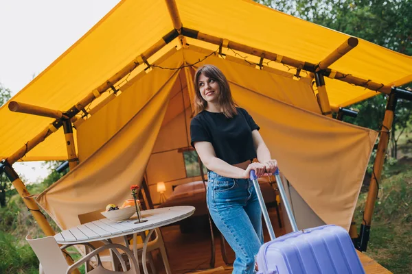 Happy woman traveler with luggage standing on background of cozy glamping house. Luxury camping tent for outdoor summer holiday and vacation. Lifestyle concept