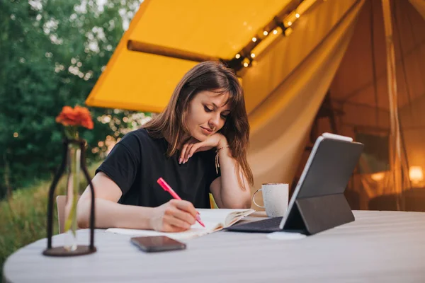 Happy Woman freelancer using laptop and making notes sitting on cozy glamping tent in a sunny day. Luxury camping tent for outdoor summer holiday and vacation. Lifestyle concept