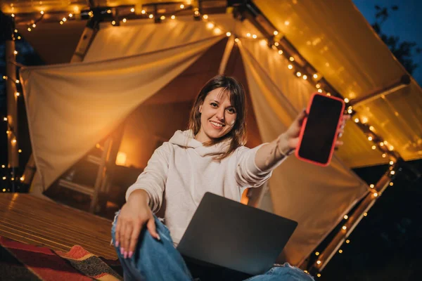 Happy Woman freelancer showing phone while working laptop on cozy glamping tent in summer night. Luxury camping tent for outdoor holiday and vacation. Lifestyle concept