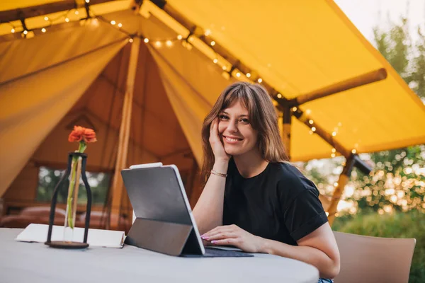 Happy Woman freelancer using a laptop on a cozy glamping tent in a sunny day. Luxury camping tent for outdoor summer holiday and vacation. Lifestyle concept