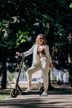 Beautiful young woman in sunglasses and white suit is standing with her electric scooter in city parkland