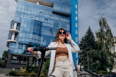Beautiful young woman in sunglasses and white suit standing on her electric scooter near modern building and looking away 