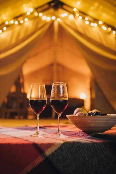 Two glass of red wine and plate with fruits standing on background of cozy glamping tent on autumn evening. Luxury camping tent for outdoor holiday and vacation. Lifestyle concept