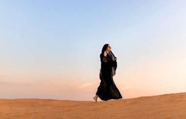 Beautiful mysterious woman in traditional arabic black long dress stands in the desert on sunset