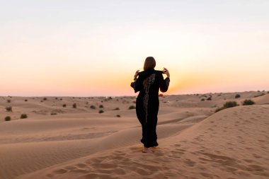 Back view of Beautiful mysterious woman in traditional arabic black long dress stands in the desert on sunset