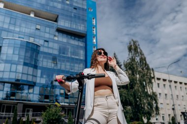 Beautiful young woman in sunglasses and white suit standing on her electric scooter near modern building and looking away 