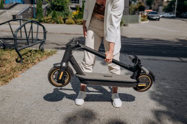 Close up of woman in white suit folding her electro scooter after ride while standing in city parkland