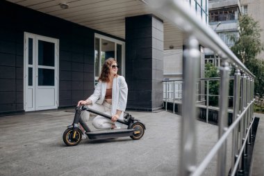 Pretty woman in white suit folding her electro scooter after ride while standing on background of modern building
