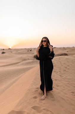 Beautiful mysterious woman in traditional arabic black long dress stands in the desert on sunset