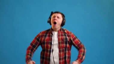 Handsome European man active dancing and jumping, listening hard rock music in headphones and enjoying sounds posing on blue studio background 