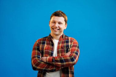 Young handsome man with happy face smiling with crossed arms looking at the camera over blue studio background. Positive person