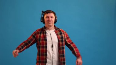 Handsome European man active dancing and jumping, listening hard rock music in headphones and enjoying sounds posing on blue studio background 