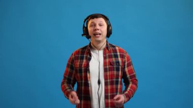 Smiling young man wearing headset freelance worker, call center or support service operator helpline, having talk with client or colleague, communication support. Young guy on blue studio background