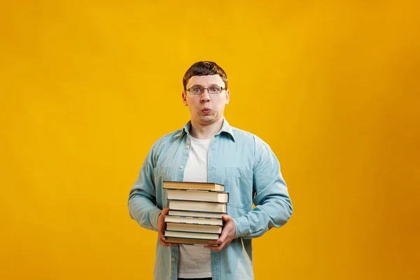 Young man student in glasses holds stack of university books from college library on yellow background. Happy guy smiles, he is happy to graduate, education abroad concept. Copy space