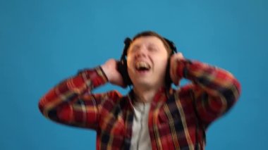 Handsome European man dancing rhythmically moving listening to music in headphones and enjoying sounds posing on blue studio background 