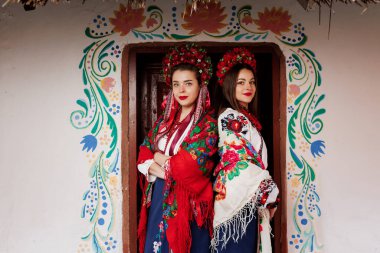 Geleneksel etnik giyimli iki Ukraynalı kadın ve süslü kulübenin arka planında çiçek kırmızısı çelenk. Vyshyvanka adında Ukrayna ulusal nakışlı bir elbise. Ukrayna için dua et.