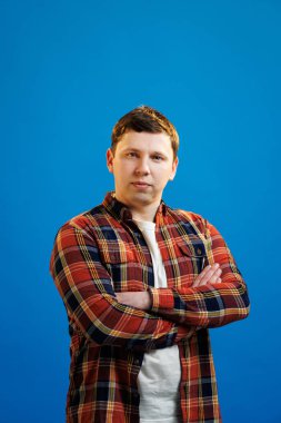 Young handsome man with focused face smiling with crossed arms looking at the camera over blue studio background. Positive person
