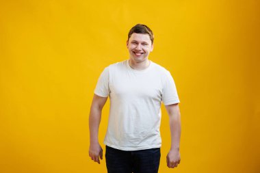 Young handsome man wearing white t-shirt with happy face smiling and looking at the camera over yellow studio background. Positive person
