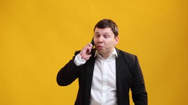 Busy businessman or manager wearing suit solves problems by talking on a mobile phone on yellow studio background