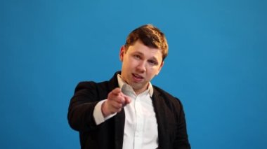 Handsome businessman in formal suit pointing to camera and looking with playful happy expression, making choice, showing direction on blue studio background. I choose you